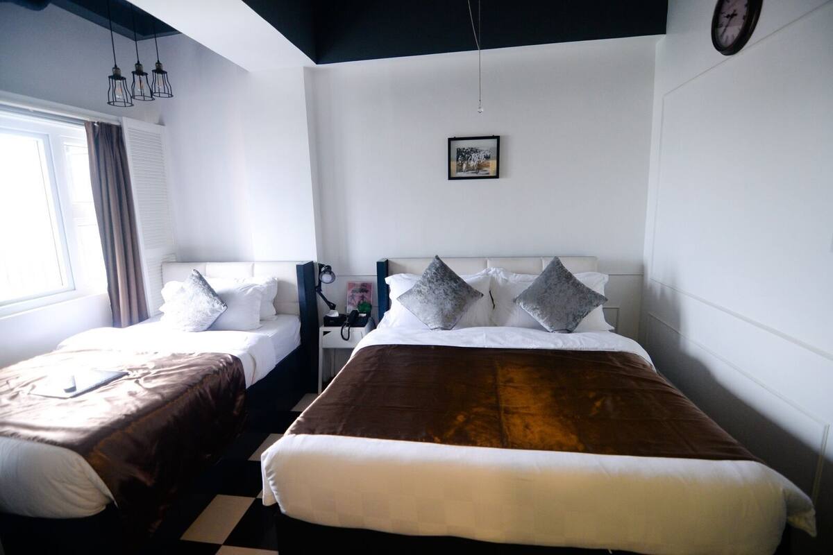 Ein Hotelzimmer mit zwei Betten, einem Schreibtisch mit Lampe, einer Wanduhr und einem Fenster mit Vorhängen.