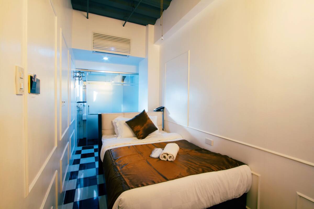 Ein Hotelzimmer mit Schachbrettmuster-Boden, einem Bett mit brauner Kopfteil, einer weißen Wand und einer sichtbaren Badezimmertür.