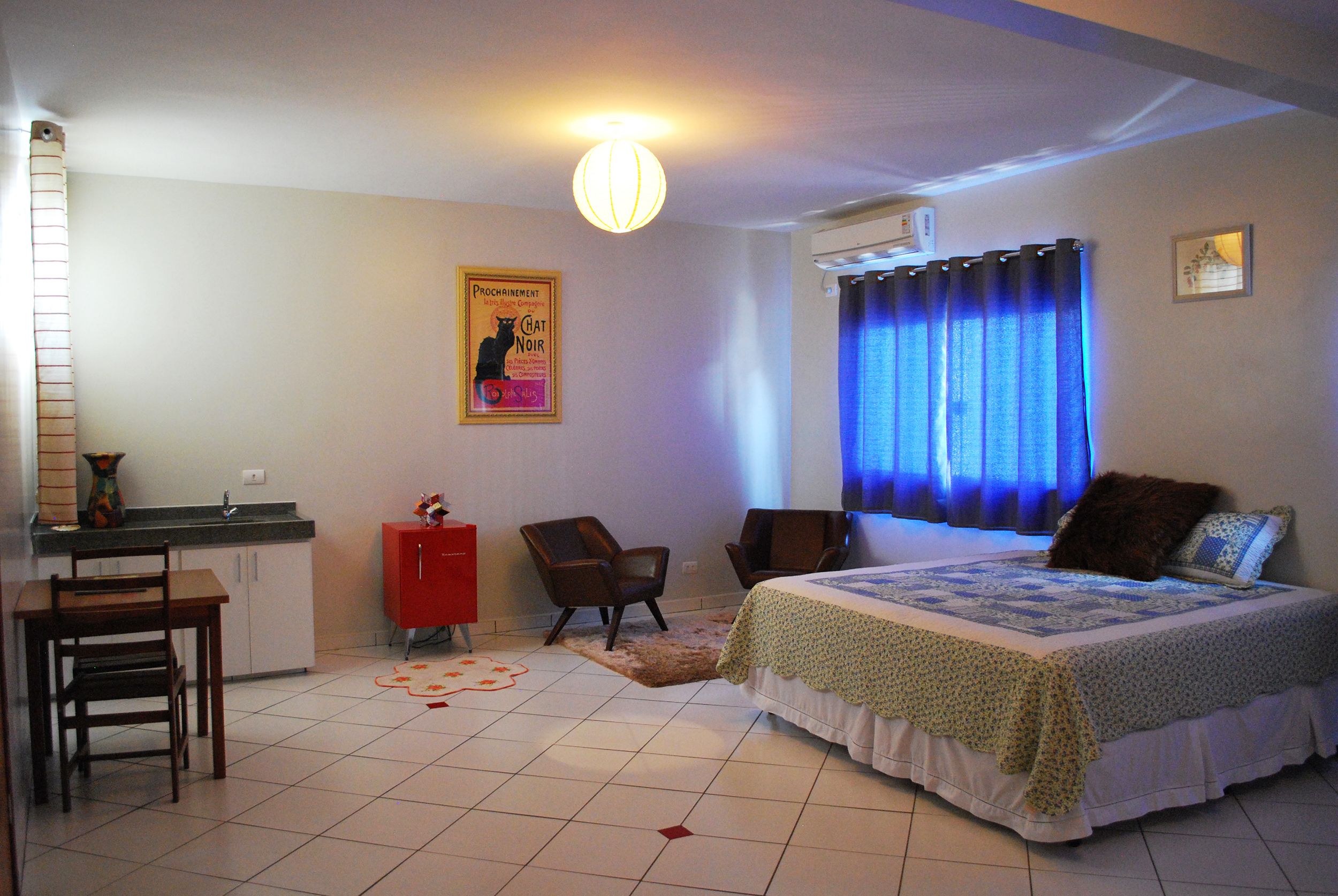 A bedroom with a bed, two chairs, a small table, a red cabinet, a poster on the wall, and a window with blue curtains.