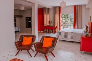 A bedroom with a bed, two chairs with orange cushions, a dining table with red curtains, and a window with a view of trees.
