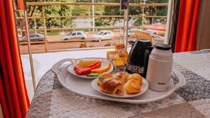 A breakfast tray with croissants, orange juice, and a water bottle on a table by a window with a view of parked cars and trees. - Pousada Beira Rio (Itumbiara)