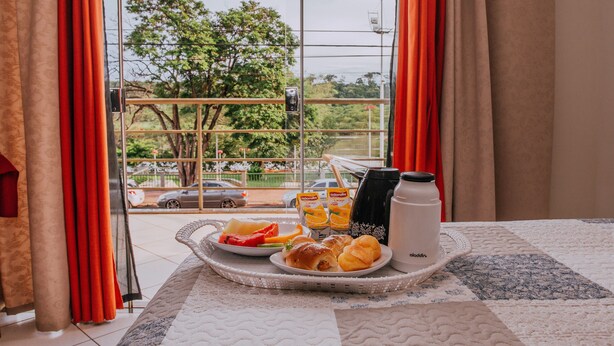A breakfast tray with croissants, fruit, and juice on a bed with a quilted bedspread, near a window with curtains and a view of trees and cars outside. - Pousada Beira Rio (Itumbiara)