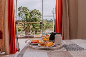 A breakfast tray with croissants, fruit, and juice on a bed with a quilted bedspread, near a window with curtains and a view of trees and cars outside.