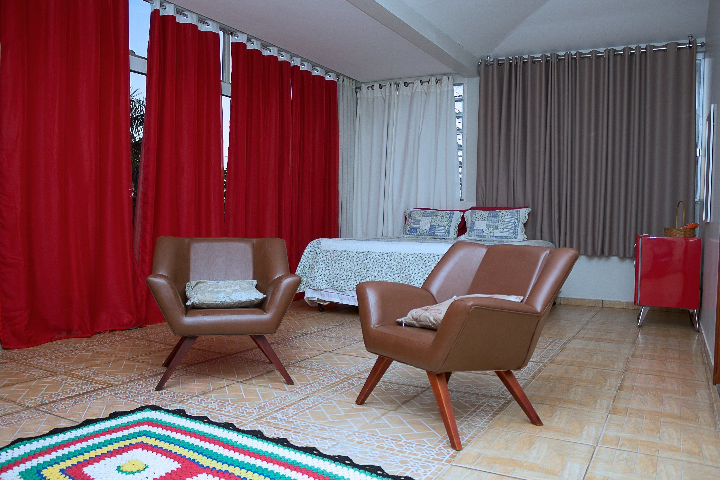 A room with two brown leather chairs, a bed with a white and blue patterned bedspread, and a red and white curtain.