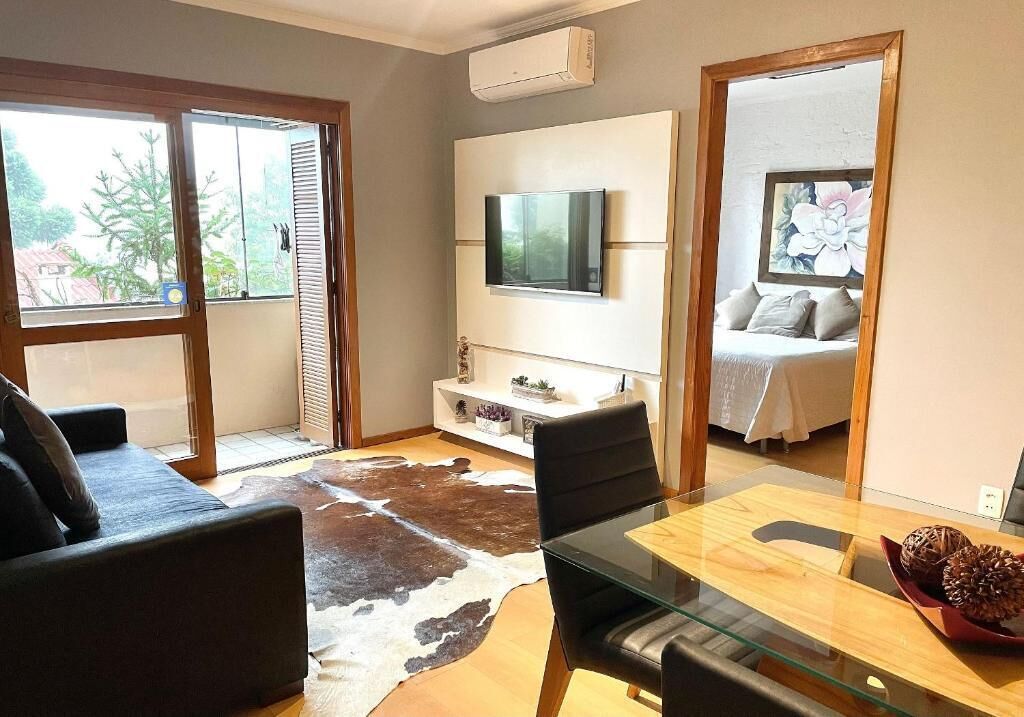 A modern living room with a glass dining table, a sofa, a TV mounted on the wall, and a bedroom visible through an open door.