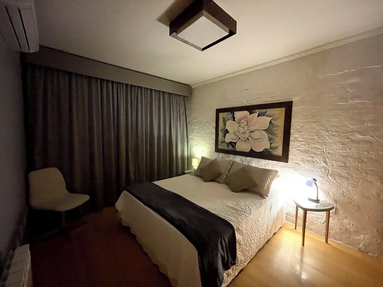 Um quarto com cama, uma cadeira, mesa de cabeceira com abajur e um quadro na parede.
