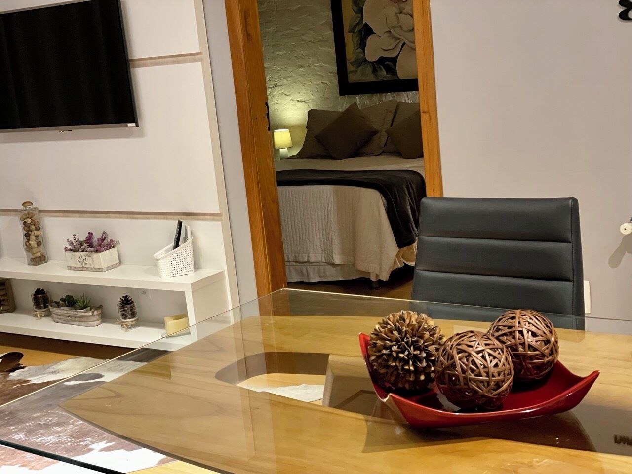 Habitació d'hotel amb una taula de vidre, un safata vermella amb boles decoratives, un llit amb edredó blanc i un televisor fixat a la paret.