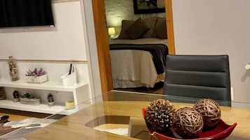 Quarto de hotel com mesa de vidro, bandeja vermelha com bolas decorativas, cama com edredão branco e televisão presa na parede.
