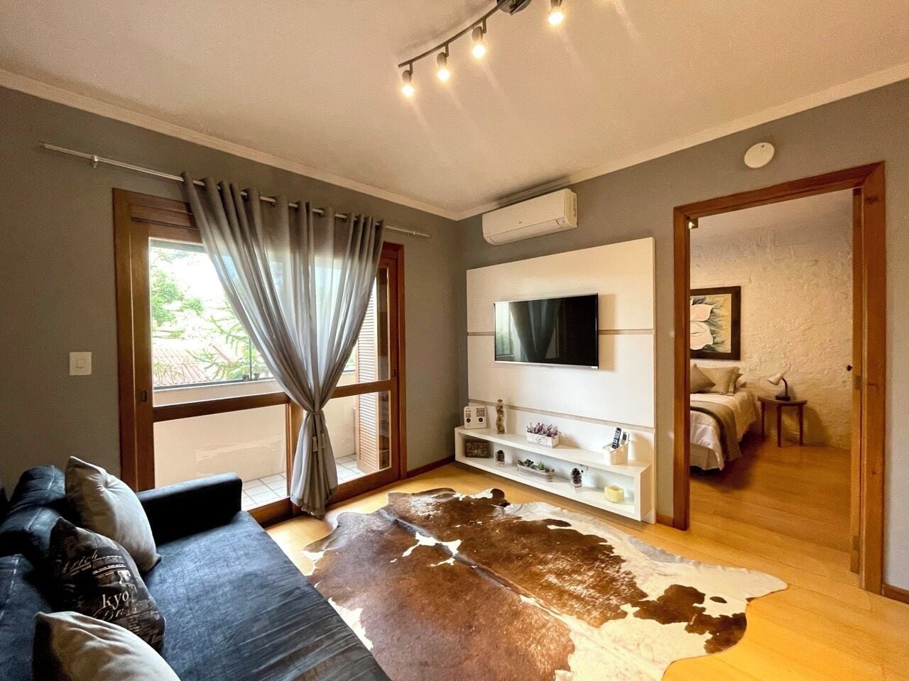 Uma sala de estar com um sofá, um tapete grande, uma televisão de tela plana e uma porta deslizante que dá para um quarto.