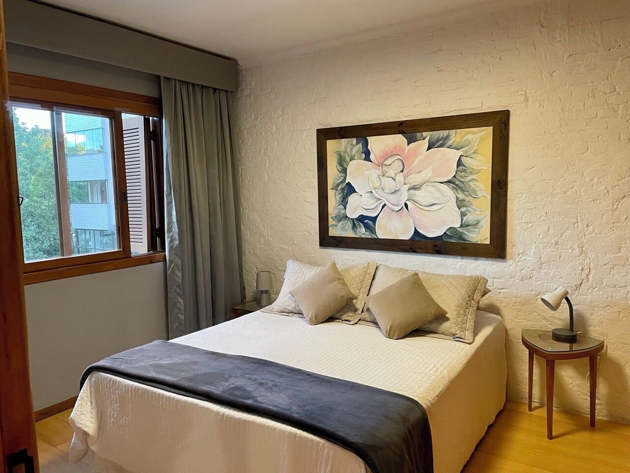 A bedroom with a bed, a nightstand, a lamp, a window with curtains, and a painting on the wall.