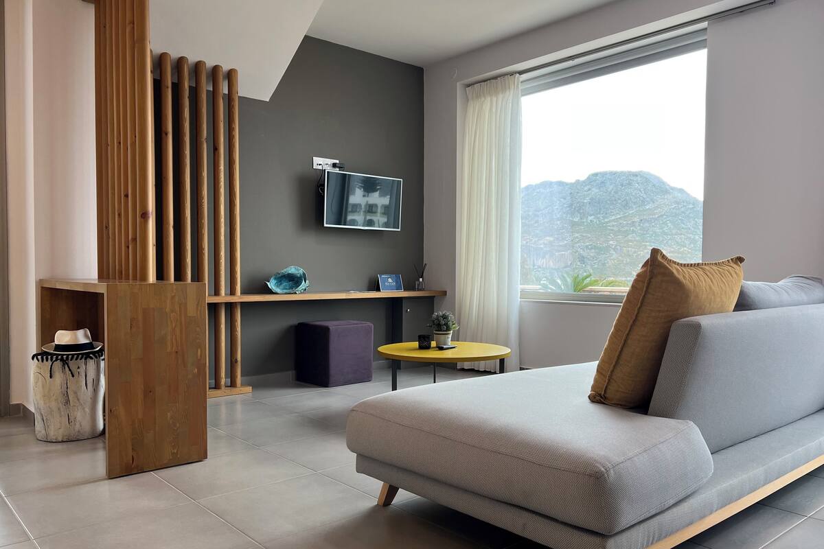 Ein modernes Wohnzimmer mit einer Couch, einem kleinen runden Tisch, einer Holzvertellung und einem großen Fenster mit Bergblick.