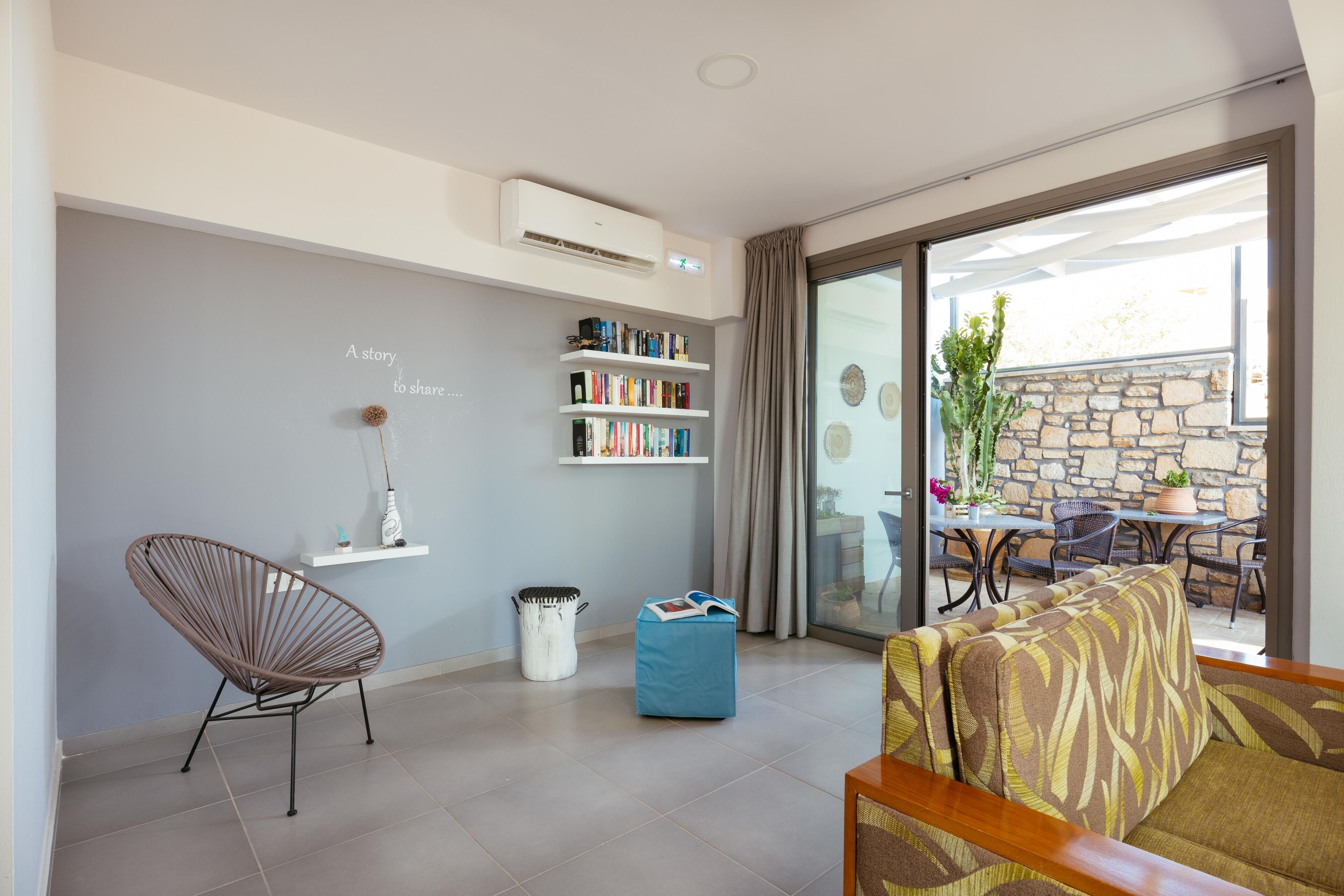 A modern living room with a grey wall, a bookshelf, a wicker chair, a blue ottoman, and a patterned sofa.