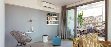 A modern living room with a grey wall, a bookshelf, a wicker chair, a blue ottoman, and a patterned sofa.