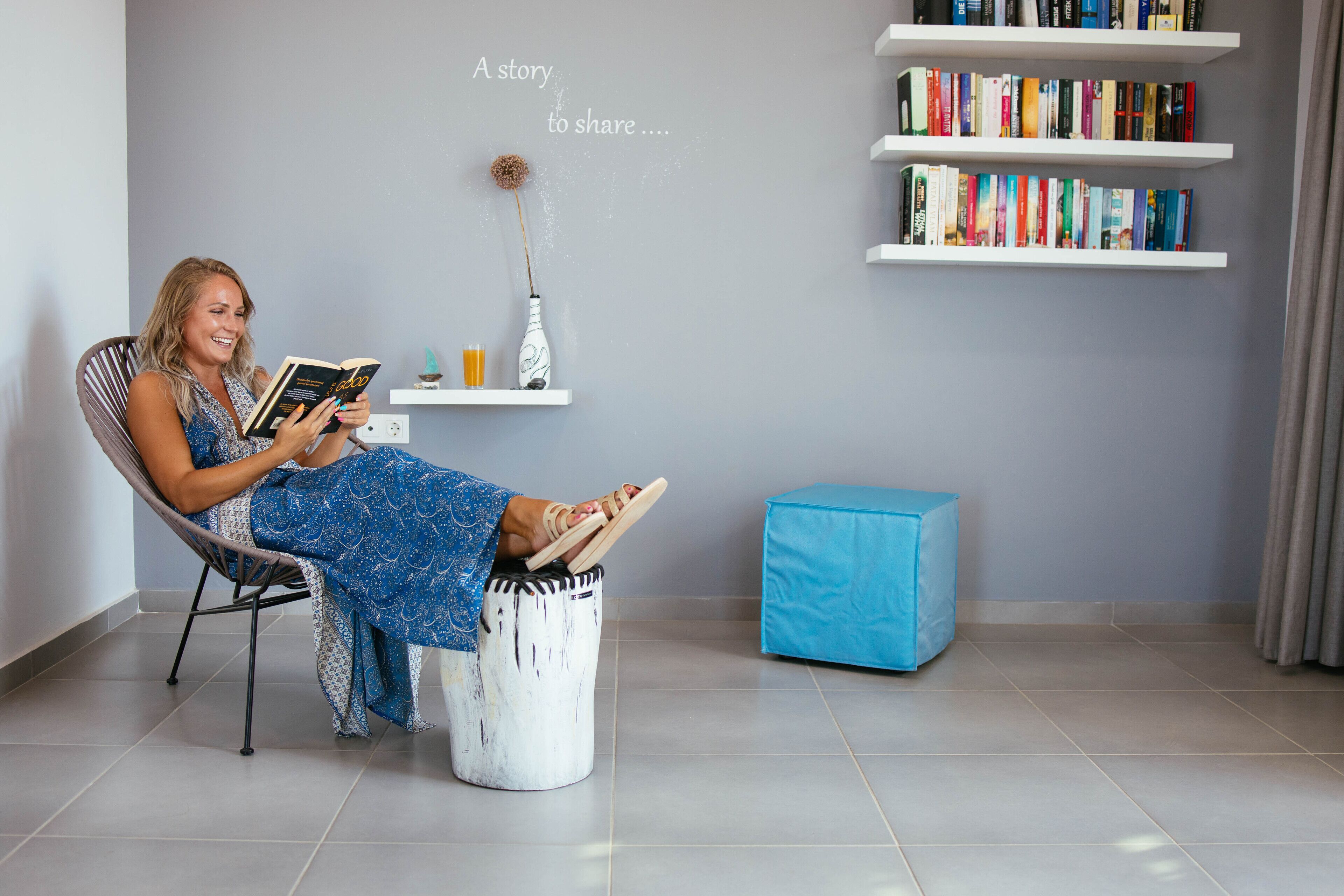 Une personne est assise dans une pièce, lisant un livre. On y voit une bibliothèque, une petite table sur laquelle est posé un vase, et un pouf bleu.
