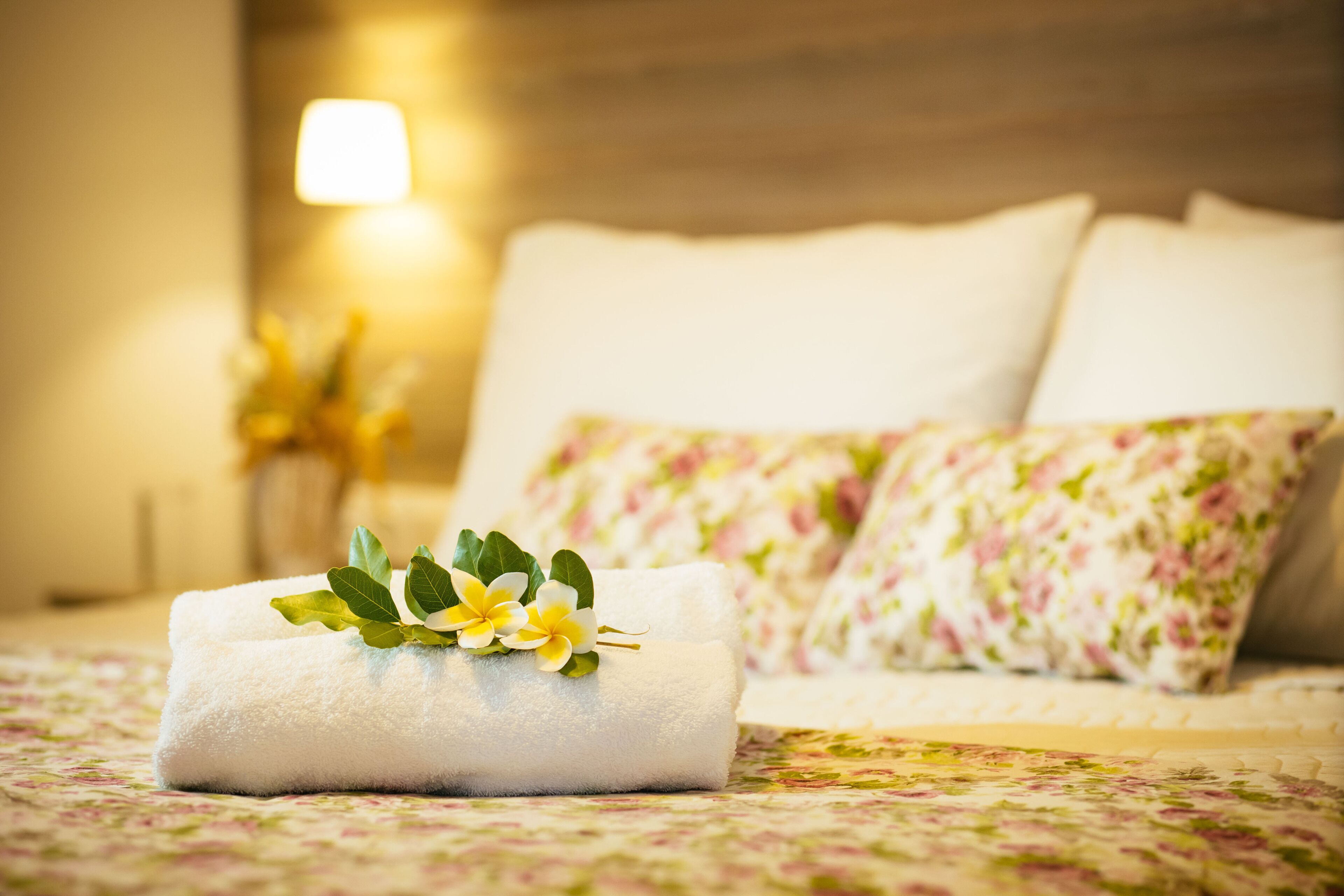 A neatly made bed with floral-patterned pillows and a rolled white towel with green leaves and yellow flowers on top.