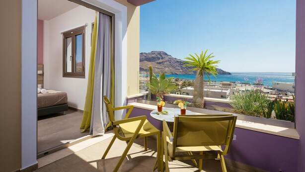 Un balcón con mesa y sillas, una cama en la habitación contigua y vistas al mar y a las montañas.