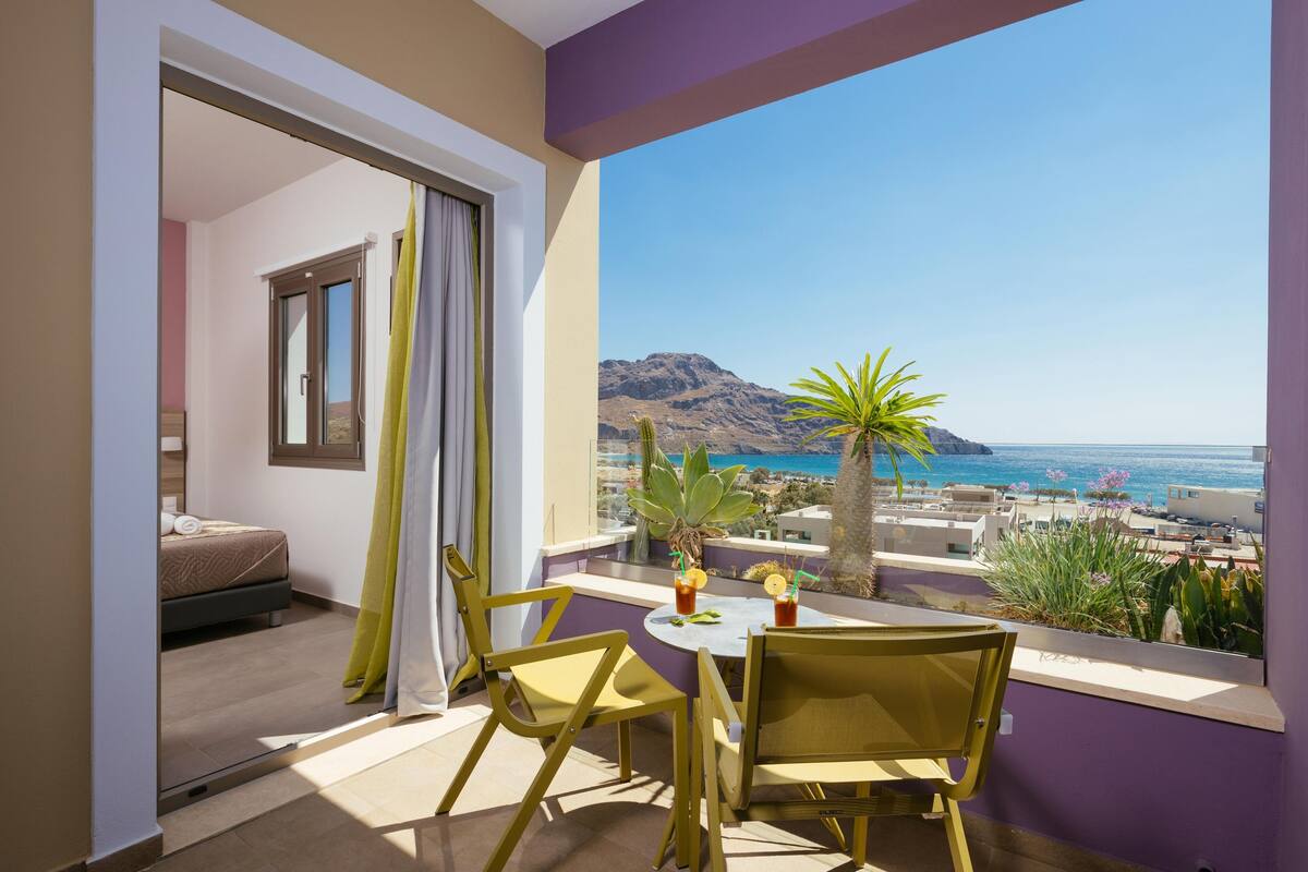 Ein Balkon mit Tisch und Stühlen, ein Bett im angrenzenden Zimmer und Blick auf Meer und Berge.