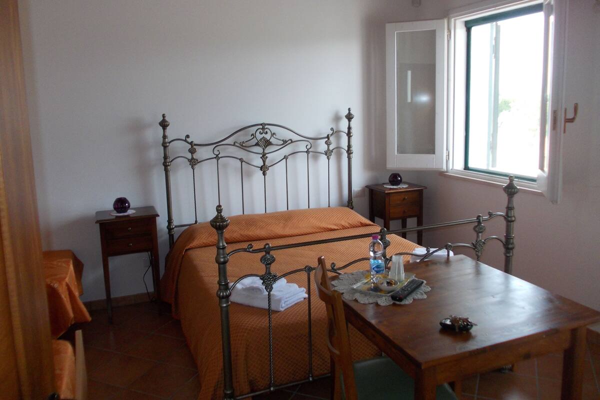Ein Schlafzimmer mit einem Metallbettgestell, einer orangefarbenen Bettdecke, einem Nachttisch aus Holz, einem Holztisch und einem Fenster mit Blick.