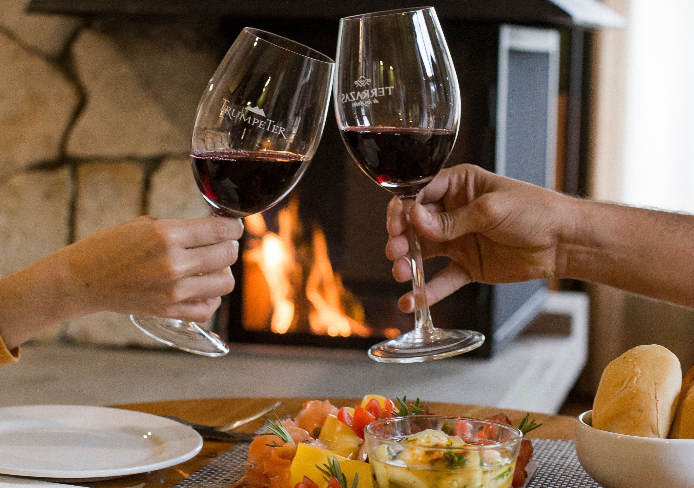 Dos personas brindando con copas de vino, una chimenea, una mesa con comida y un cuenco de pan.