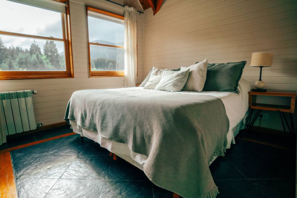 A bedroom with a bed, a window, a radiator, and a nightstand with a lamp.