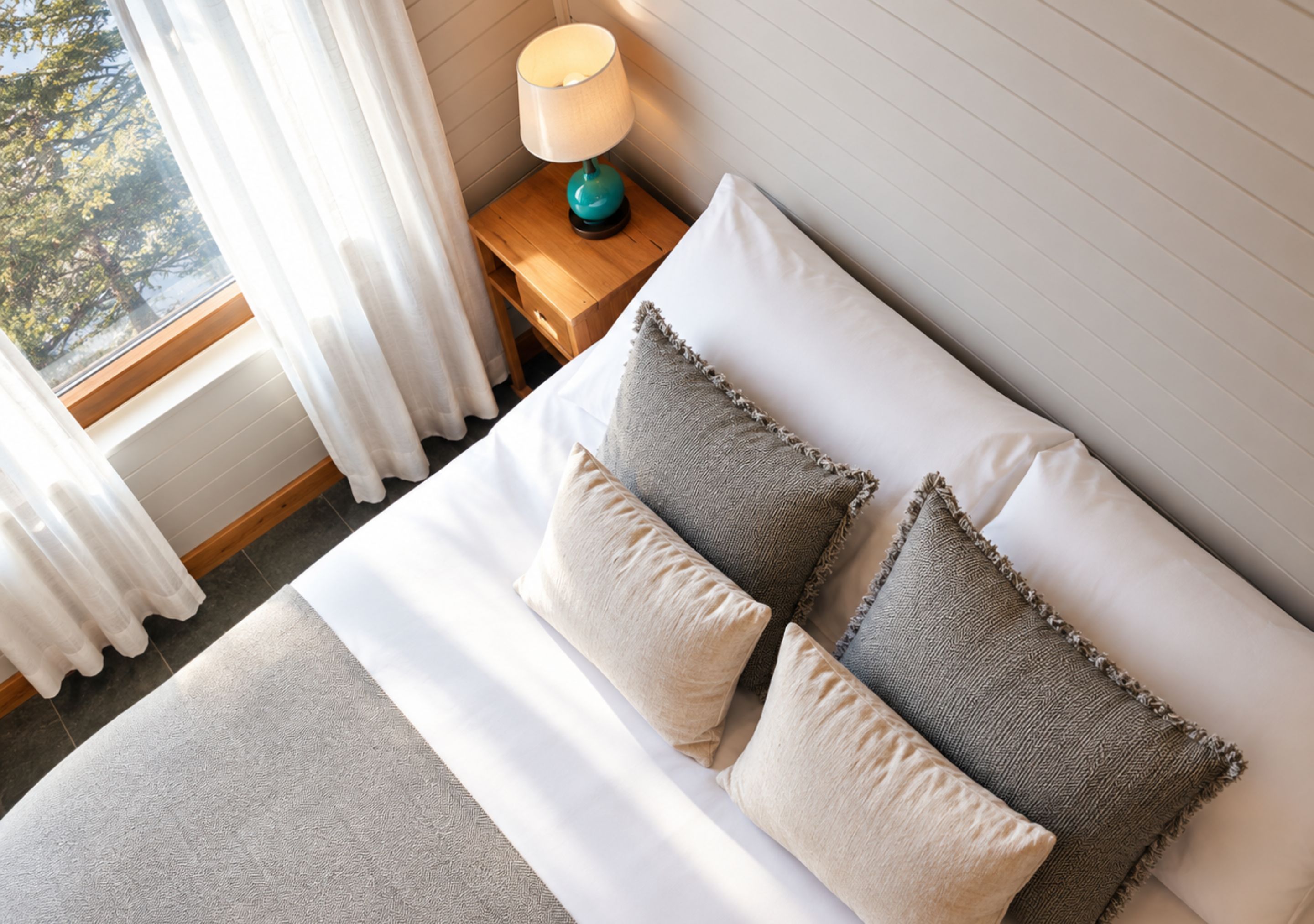 Ein ordentlich bezogenes Bett mit weißen Leinentüchern und zwei Kissen, ein Nachttisch mit einer Lampe und ein Fenster mit Vorhängen.
