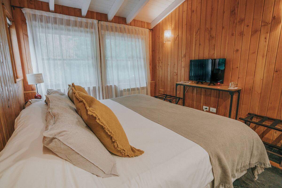 A room with wooden paneling, a bed with a beige blanket, a television on a wooden stand, and a lamp on a bedside table.