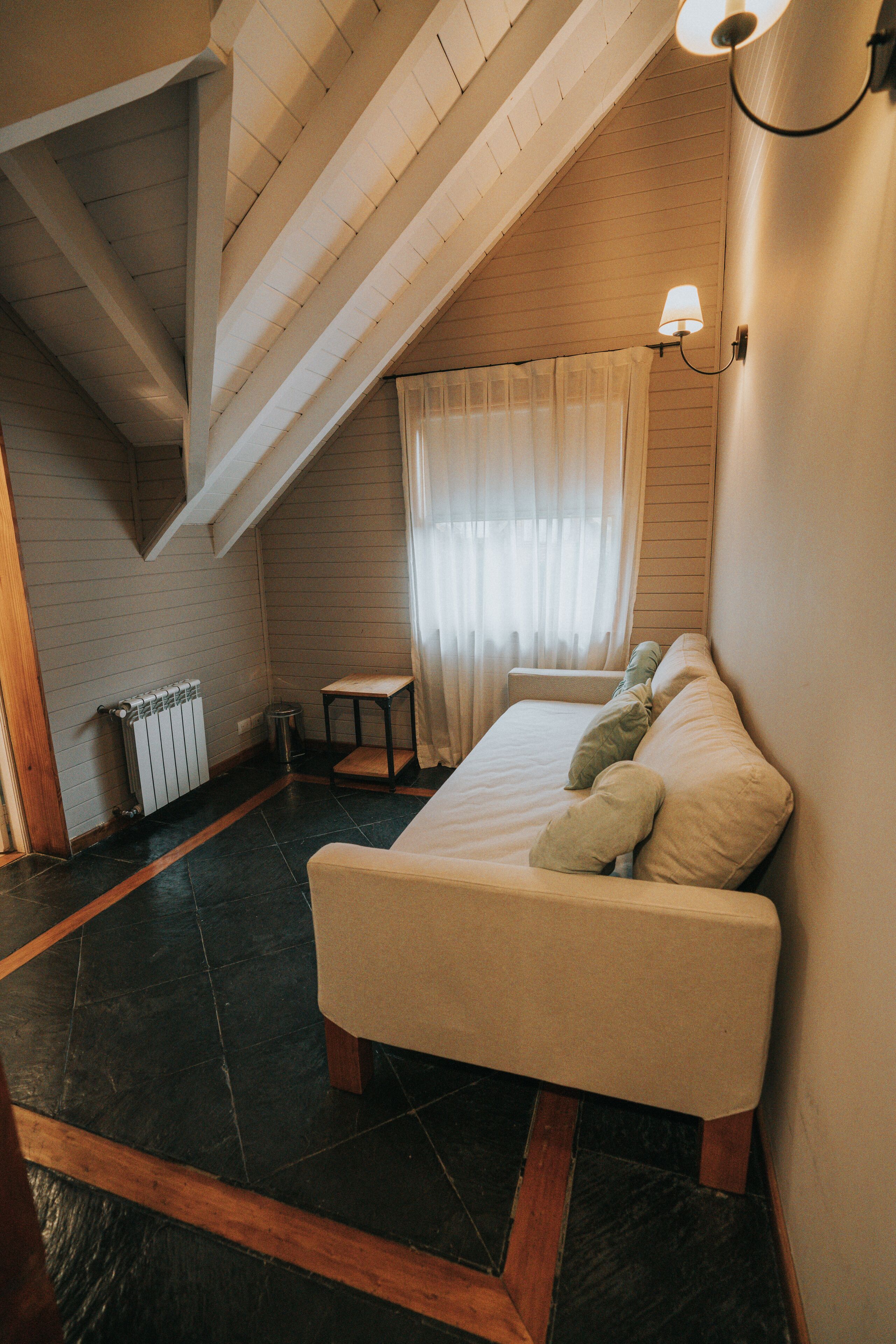 Un dormitorio con techo inclinado, una cama con almohadas, una mesita y una ventana con cortinas.