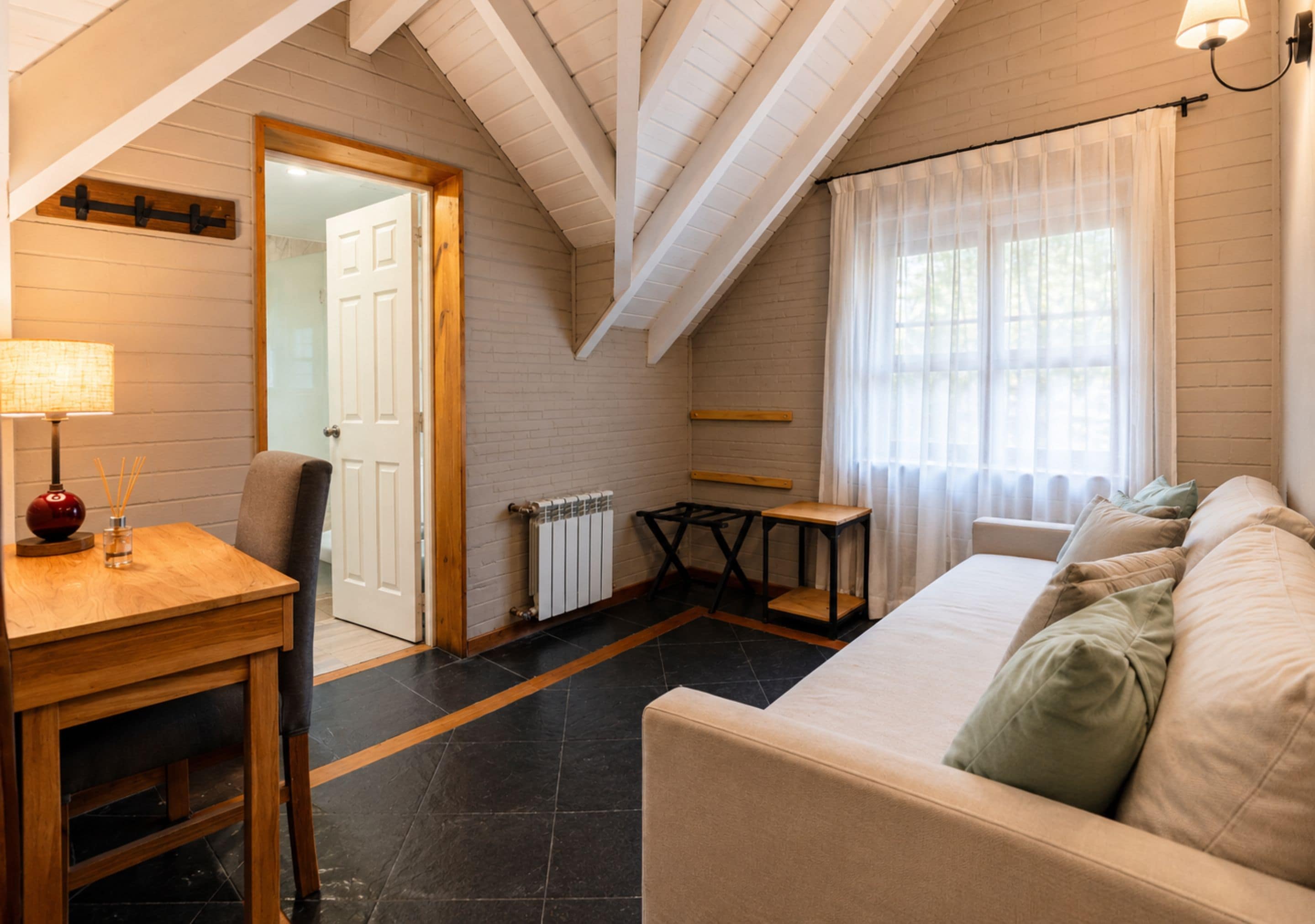 A cozy room with a wooden table, a chair, a sofa, a radiator, and a window with curtains.