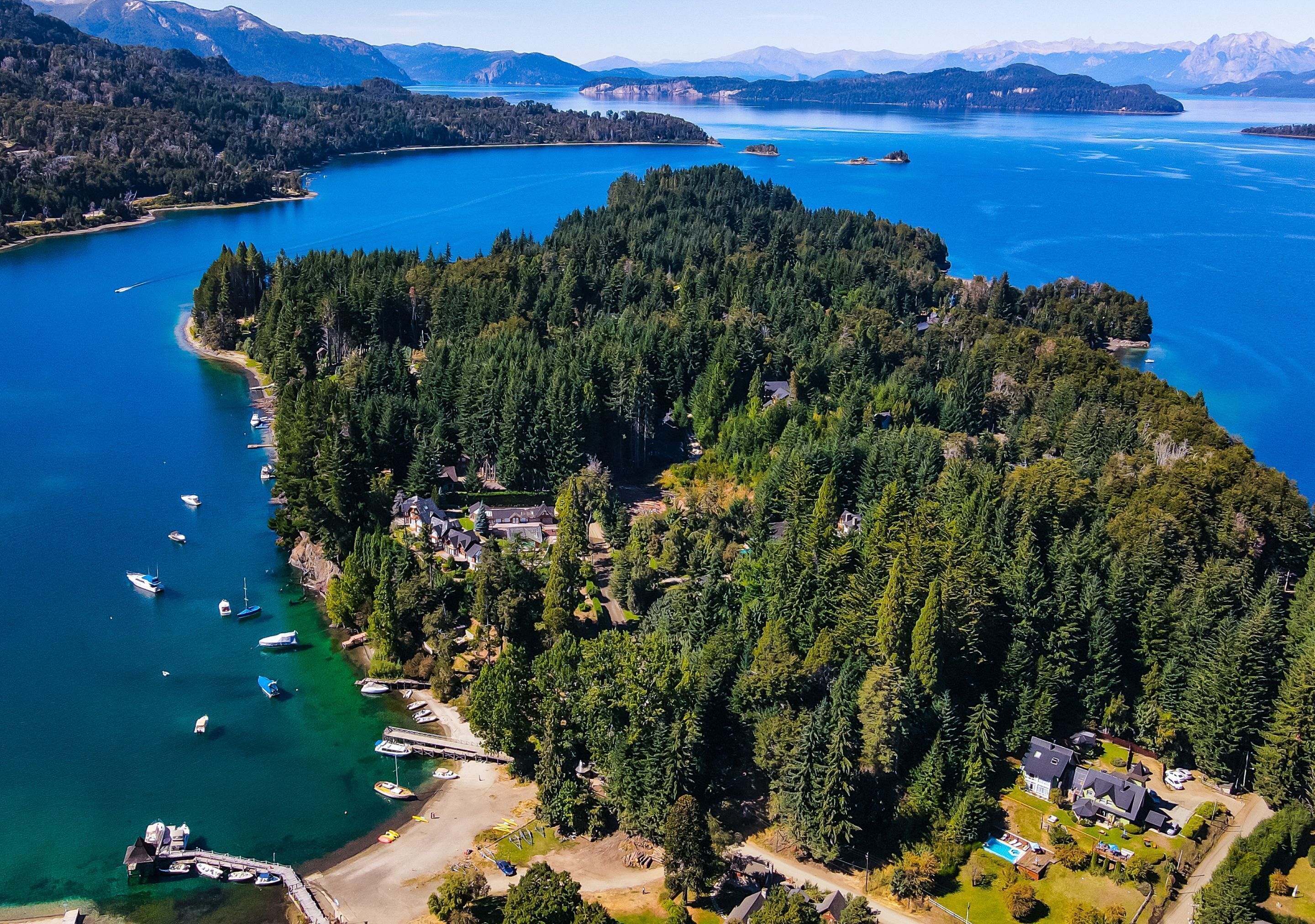 Vue aérienne d’une île boisée, avec un quai et des bateaux, entourée d’une vaste étendue d’eau et de montagnes.