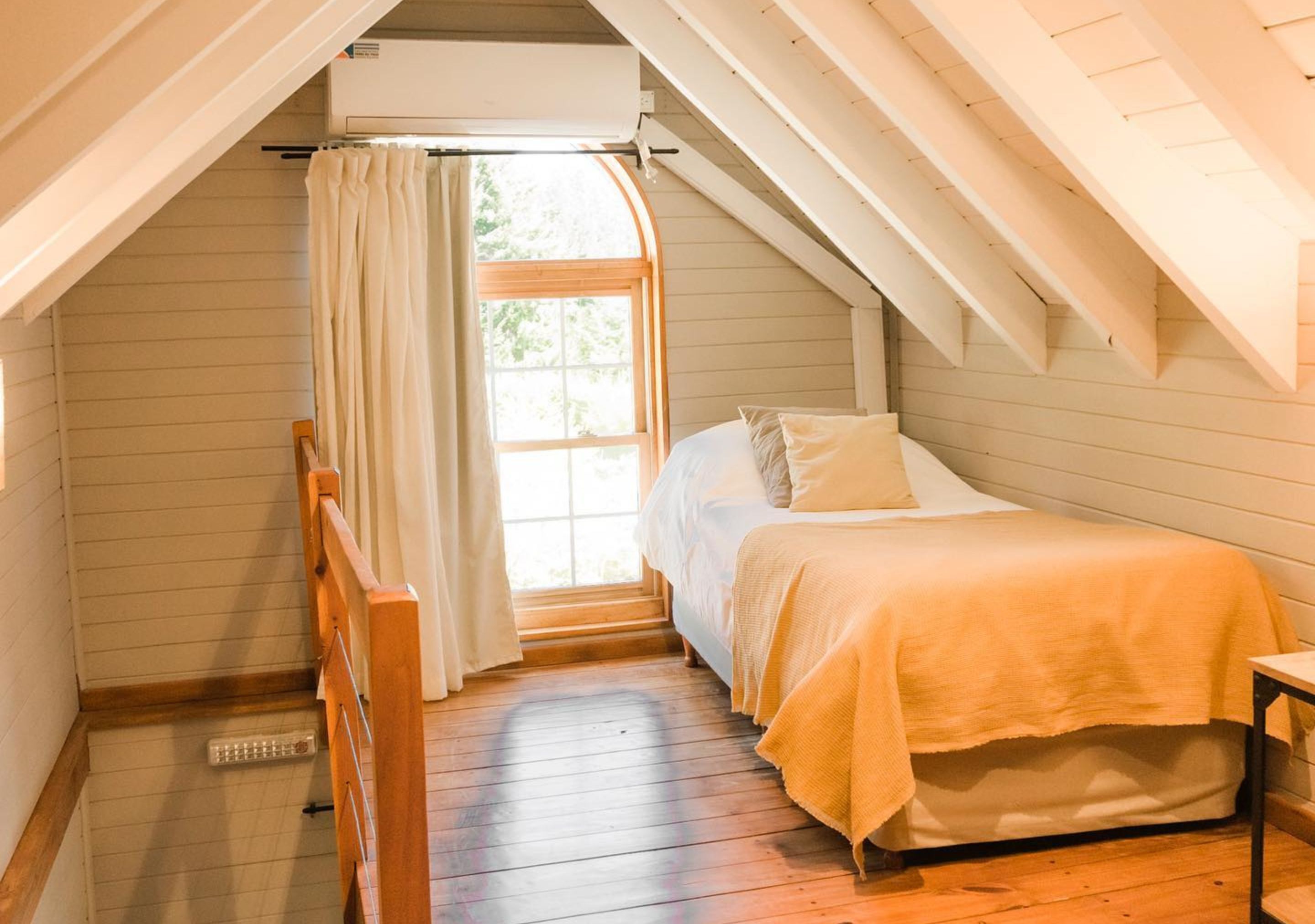 A bedroom with a wooden floor, a bed with a white and beige bedspread, a window with curtains, and a radiator.