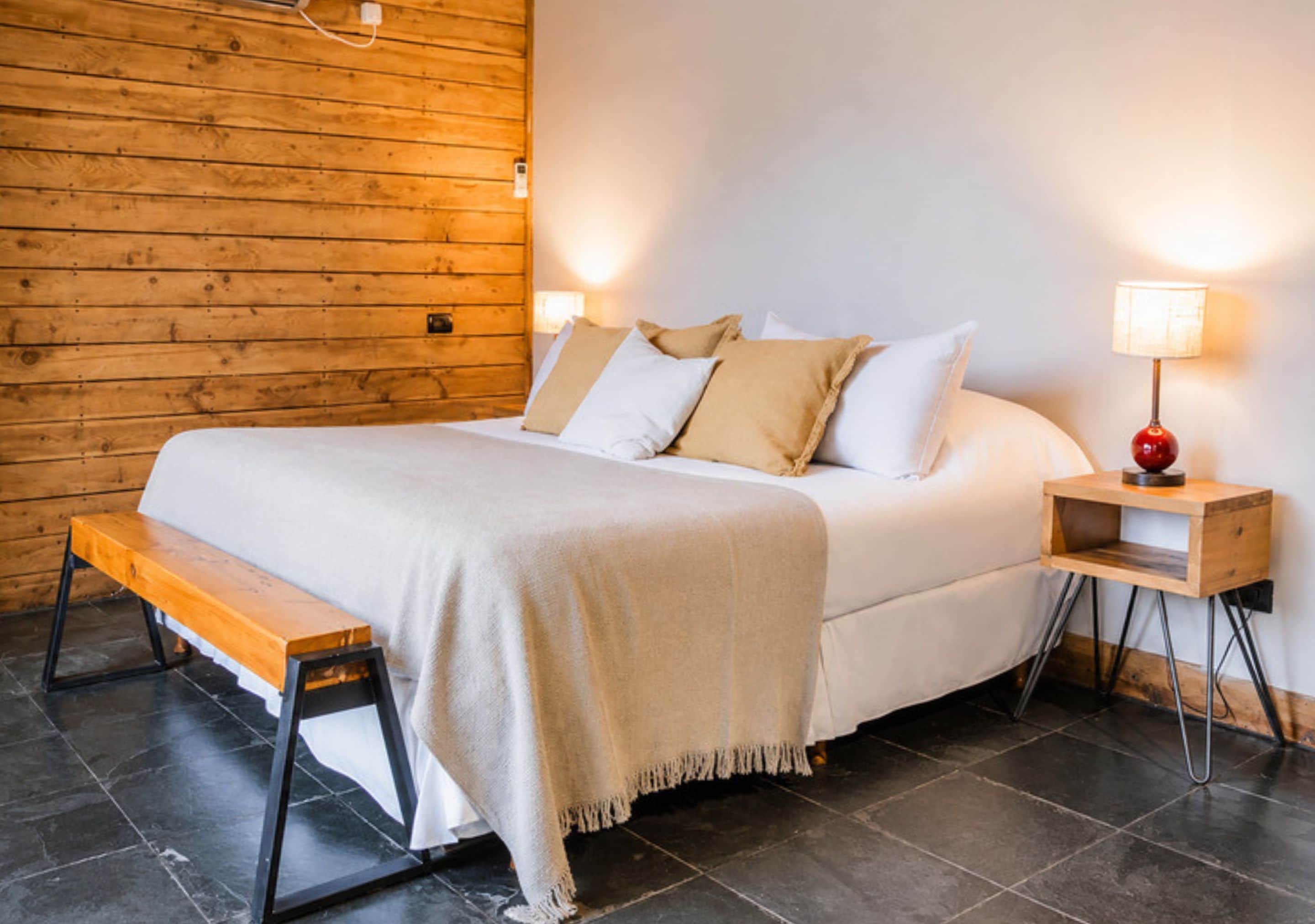 A hotel room with a wooden wall, a bed with white bedding, a bench, a nightstand with a lamp, and a tiled floor.