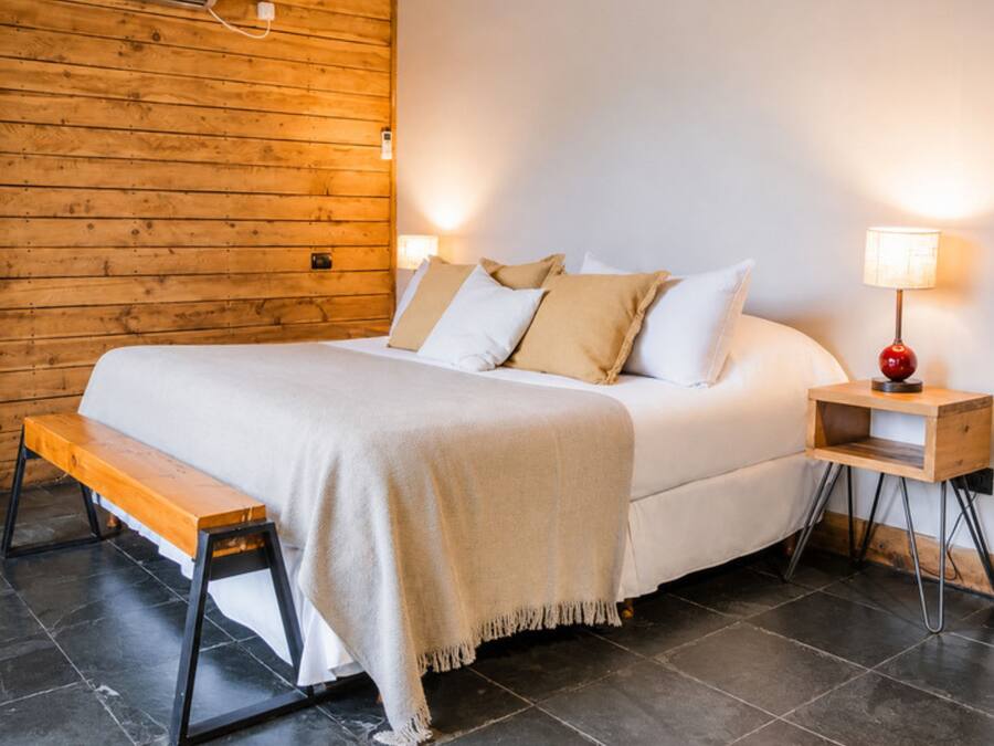 A hotel room with a wooden wall, a bed with white bedding, a bench, a nightstand with a lamp, and a tiled floor.