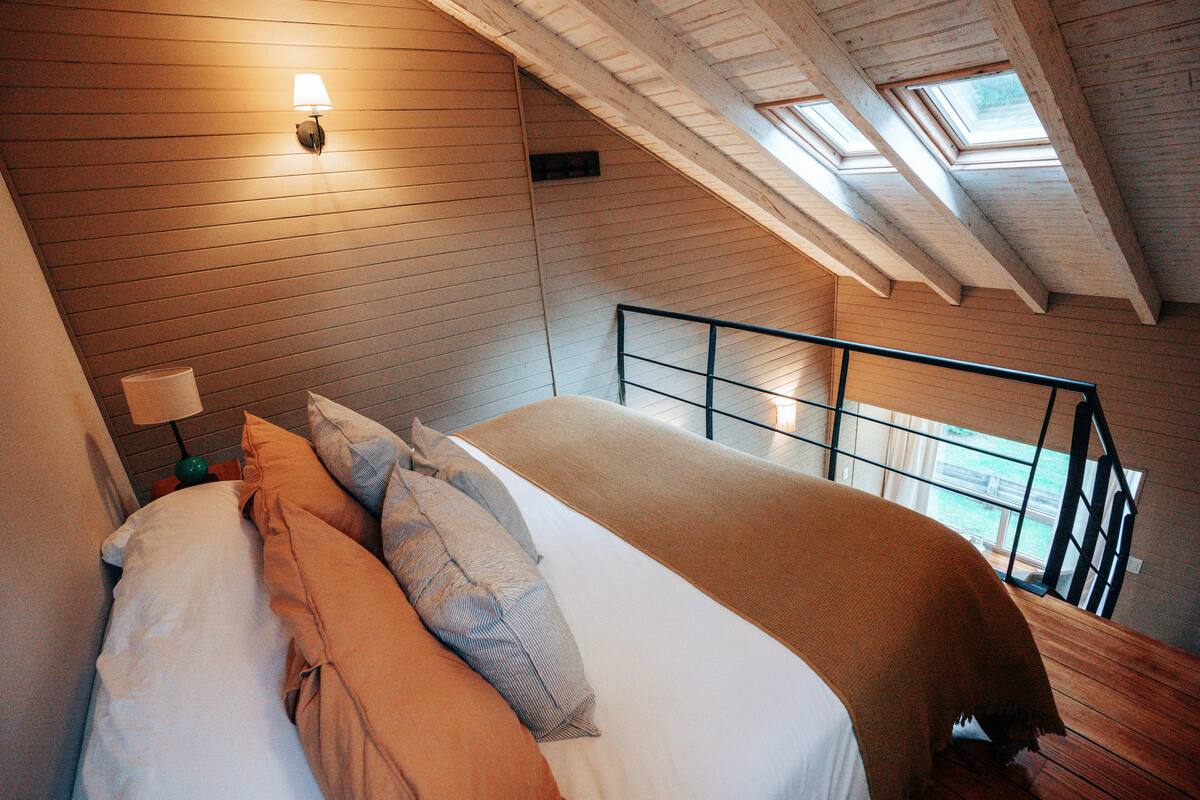 A bedroom with a bed, pillows, a nightstand, a lamp, and a skylight.