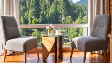 A room with two grey upholstered chairs, a glass-top table with a vase of flowers, and a view of a forested mountain range through a window.