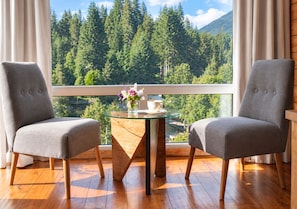 A room with two grey upholstered chairs, a glass-top table with a vase of flowers, and a view of a forested mountain range through a window. - Hosteria Verde Morada (Villa La Angostura)