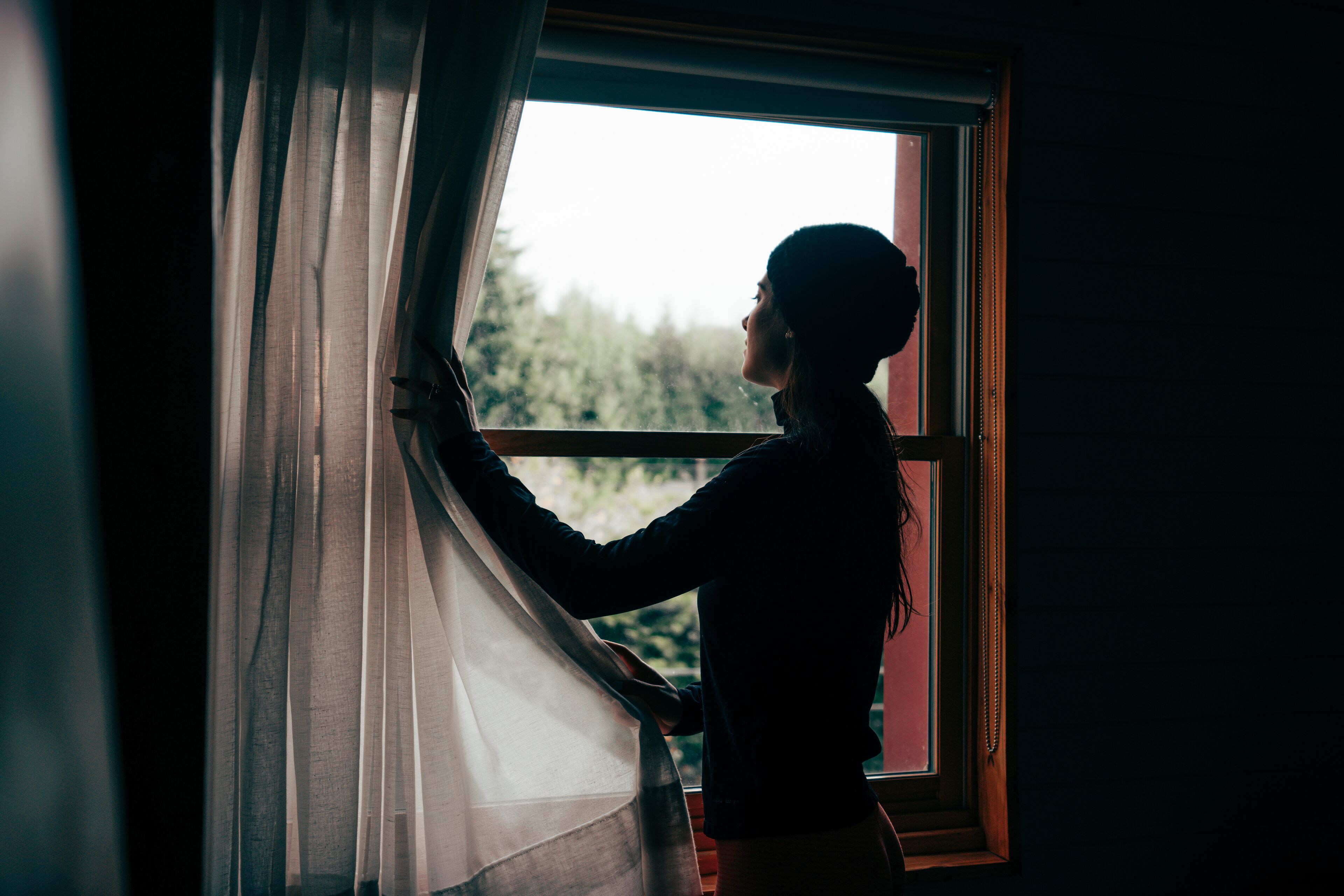 A person standing by a window with curtains, looking outside.