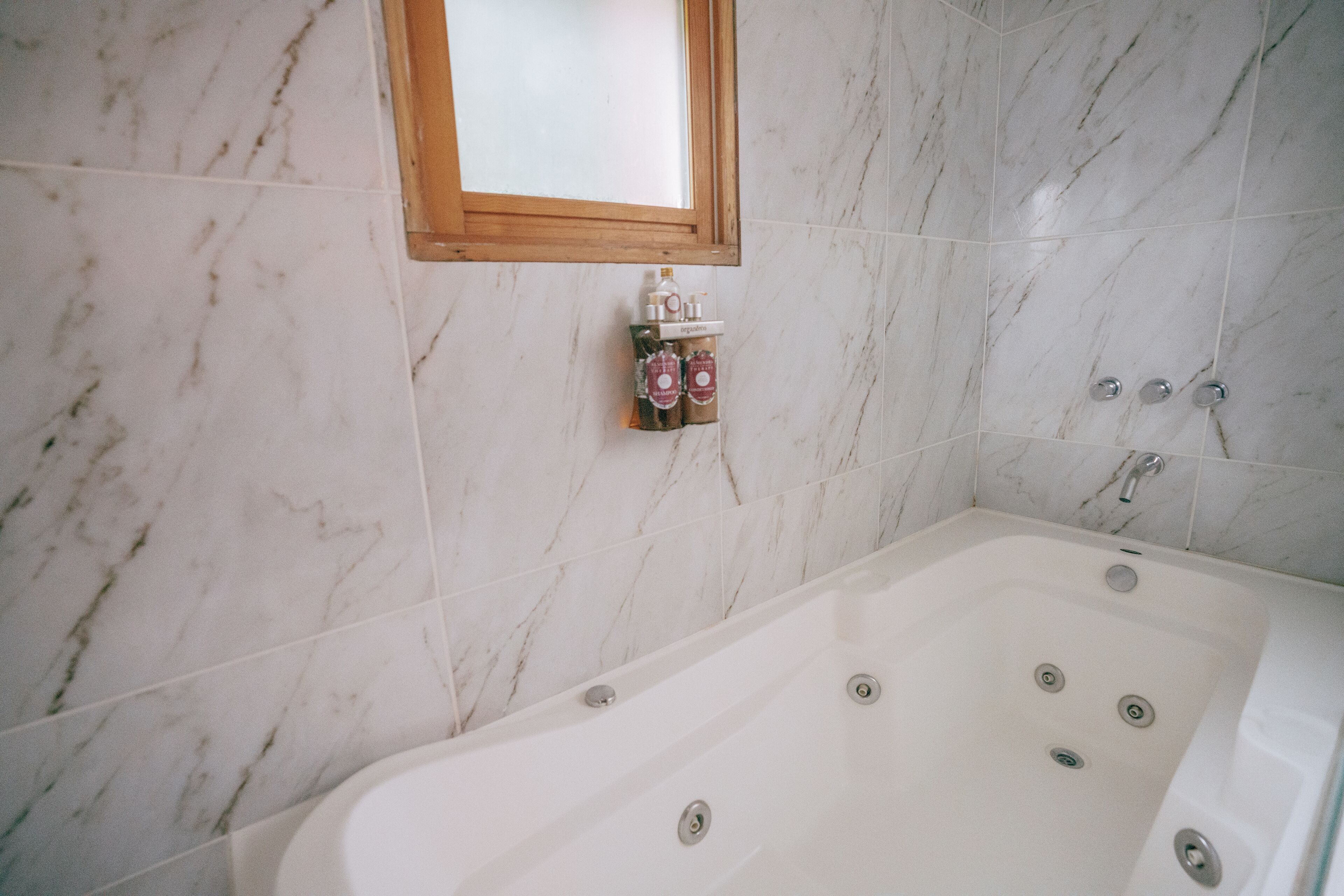 A bathroom with a marble floor, a bathtub with multiple faucets, and a small shelf with toiletries.