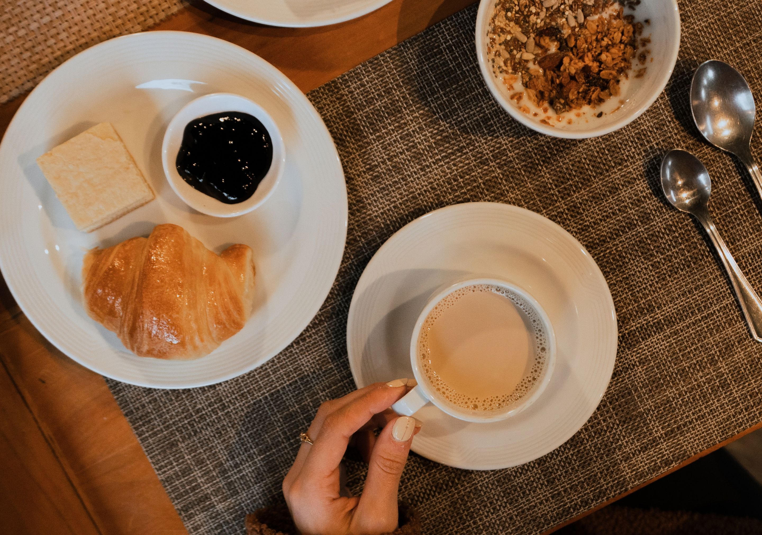 Egy asztal, rajta egy tányérnyi croissant, egy tál lekvár, egy csésze kávé és egy tál gabonapelyhes.
