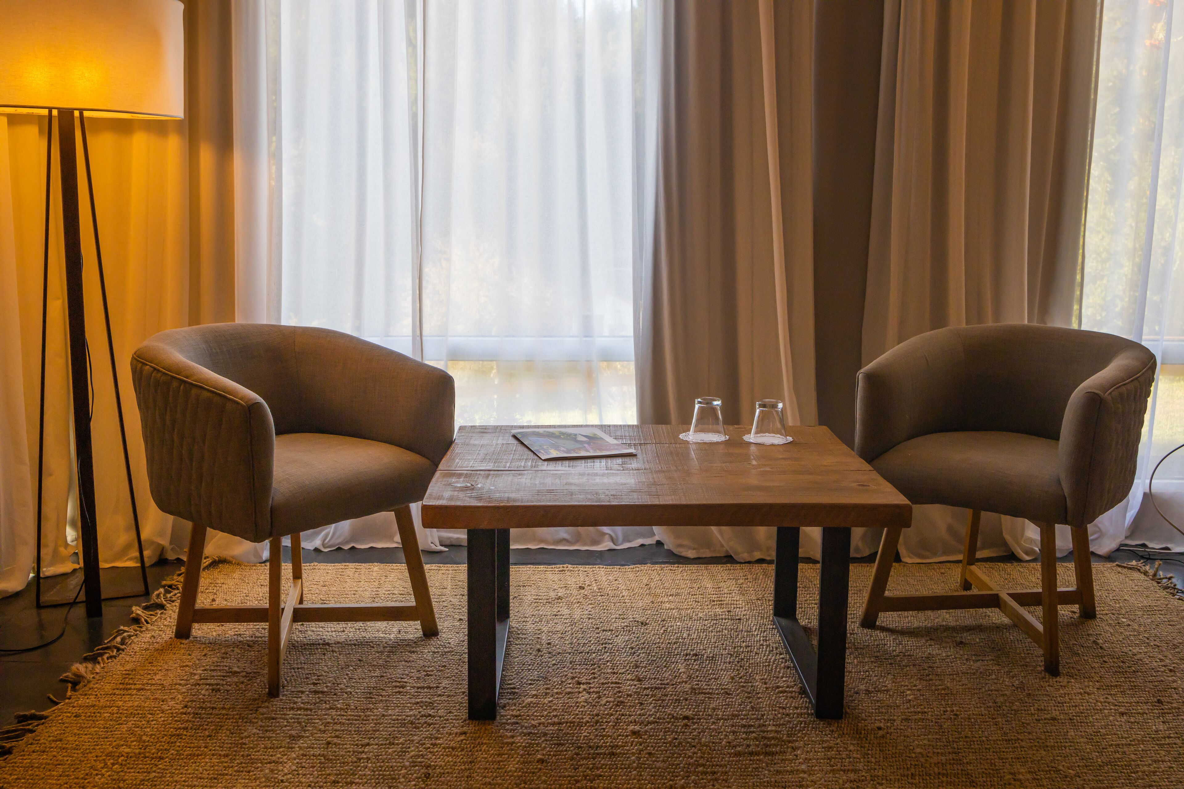 A room with two armchairs, a wooden table, a floor lamp, and a view through sheer curtains.
