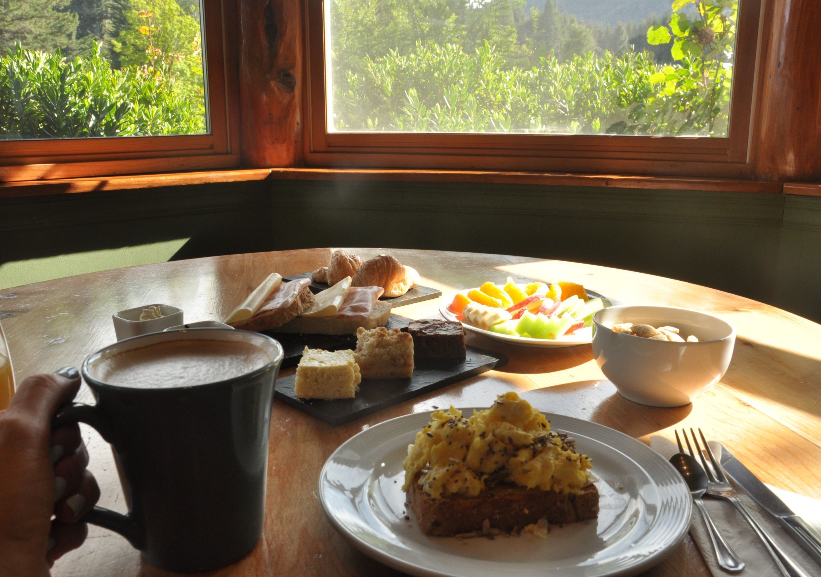A table with food and a coffee mug, a window with a view of greenery, and a wooden table.
