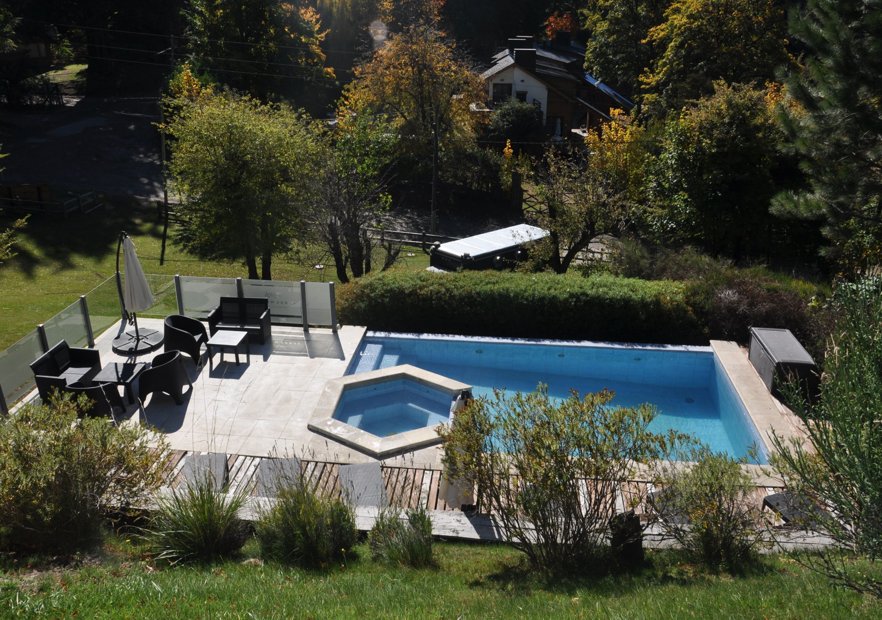A swimming pool with a deck, surrounded by greenery and a house in the background.
