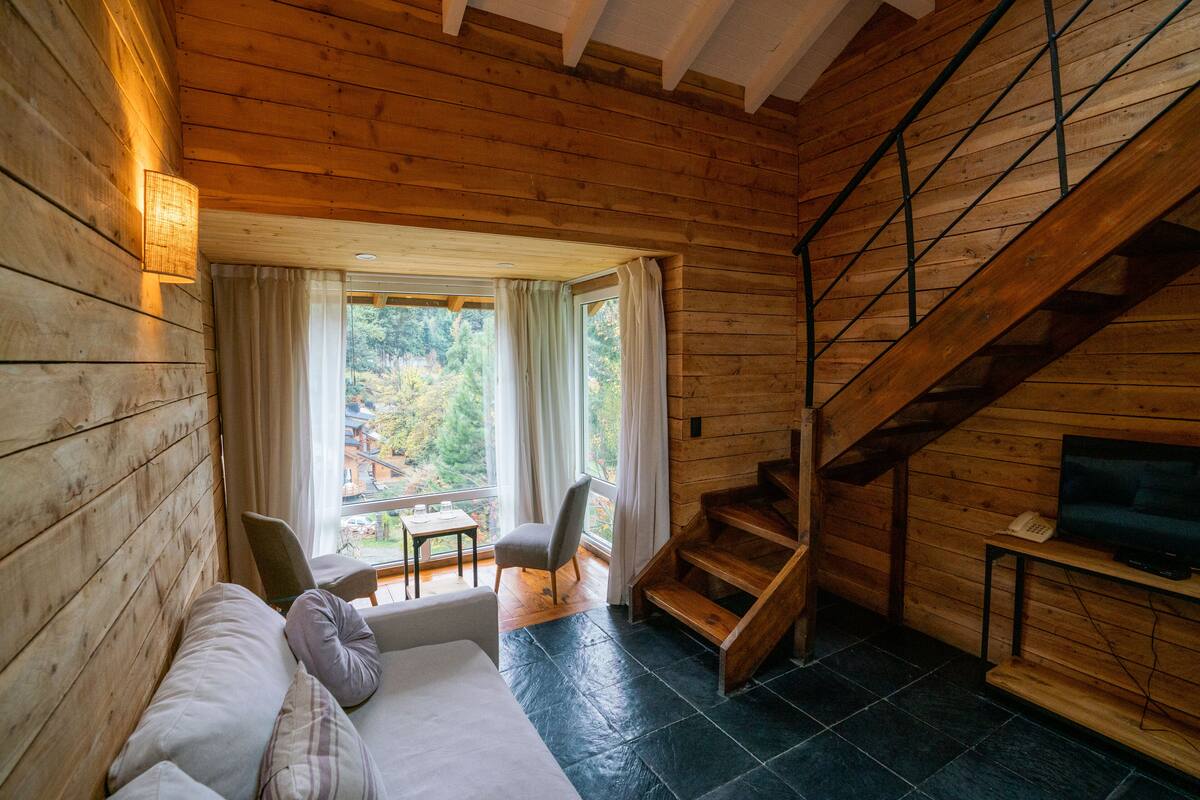 A wooden cabin interior with a sofa, a chair, a table, a staircase, and a TV.
