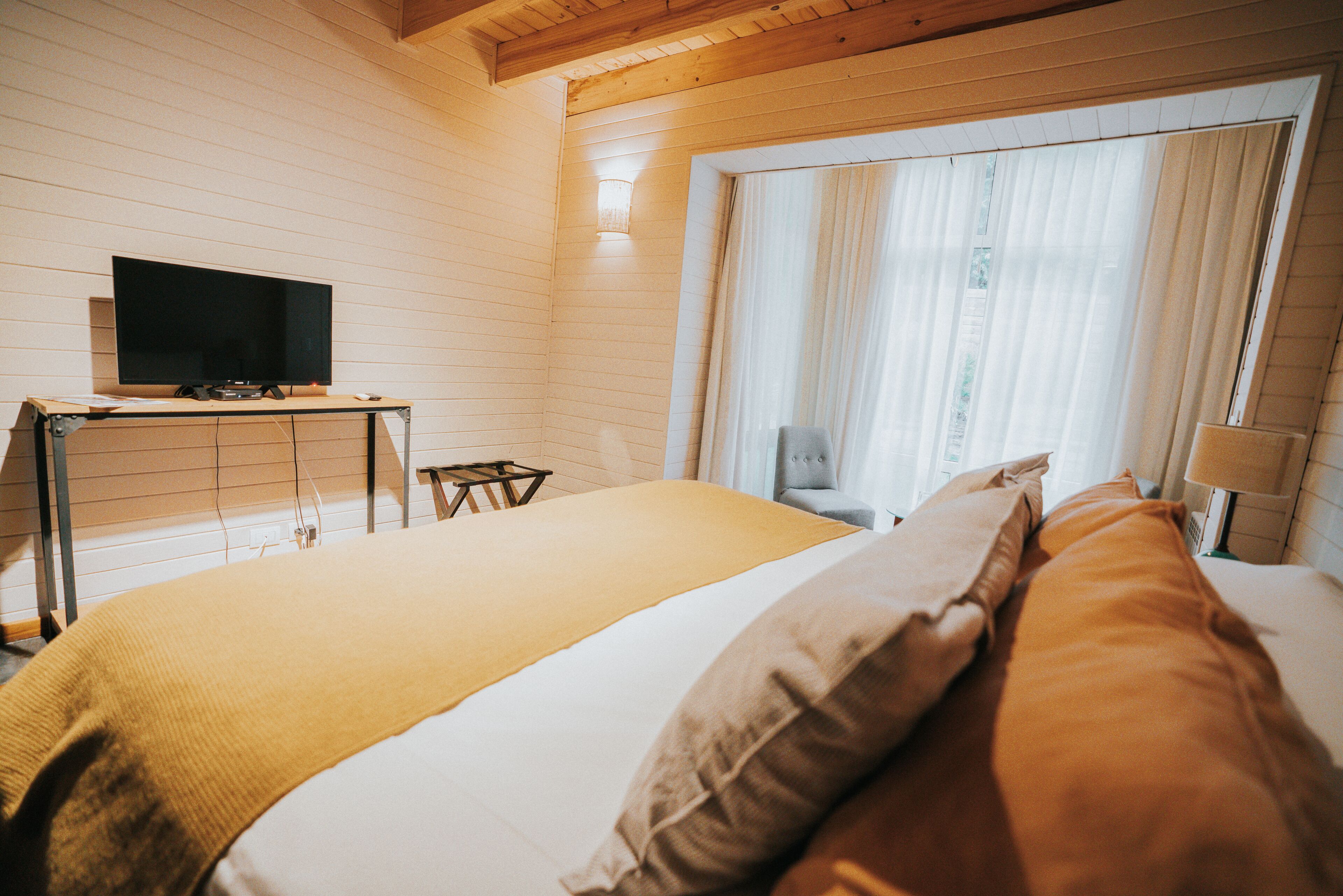 Un dormitorio con una cama, un televisor sobre un mueble bajo, una silla y una ventana con cortinas.