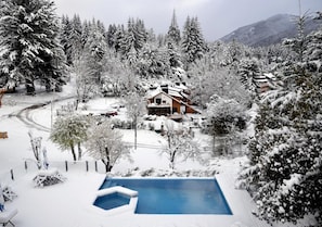 A snow-covered landscape with a swimming pool, a wooden house, and surrounding trees.