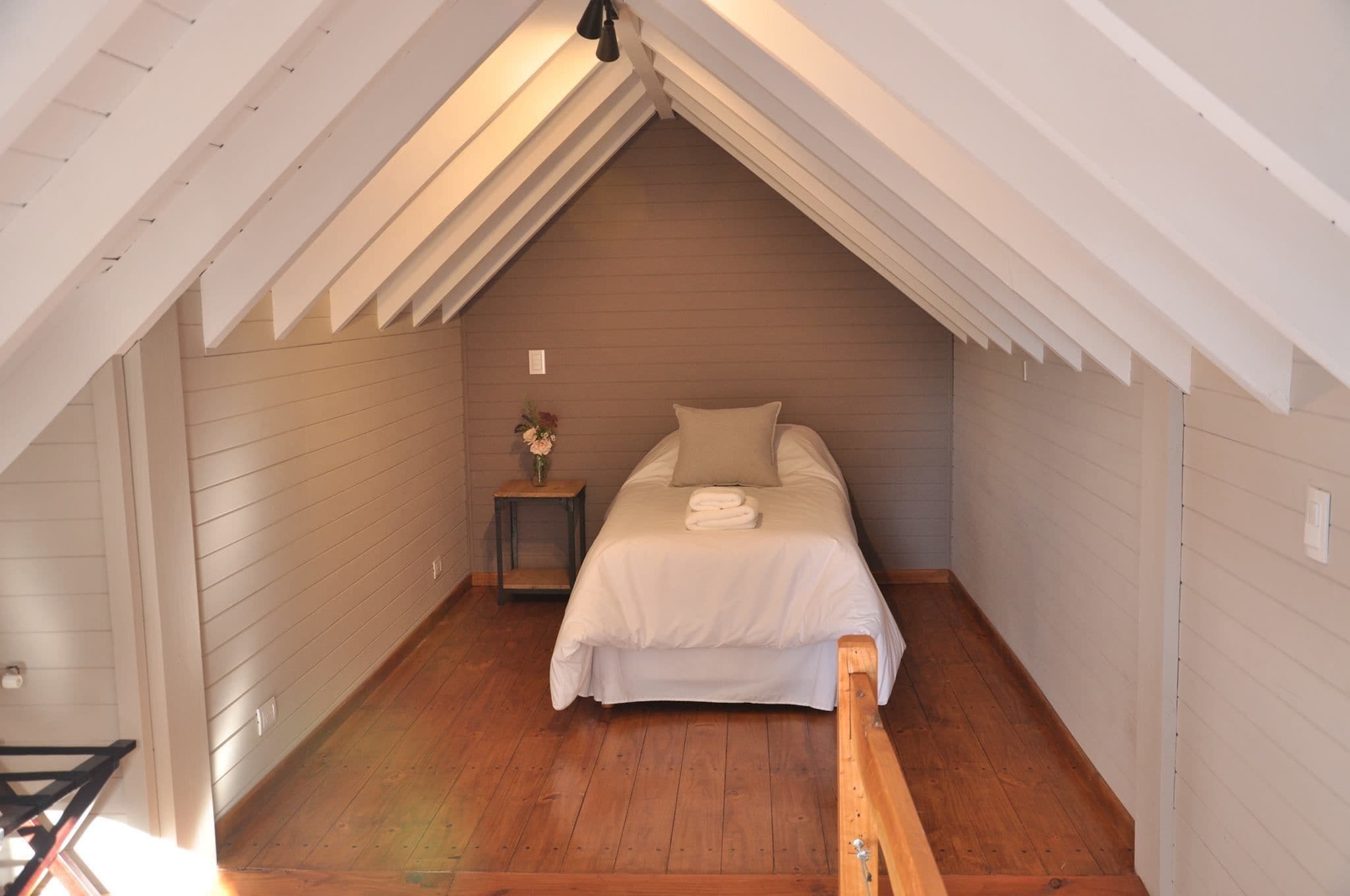 Un dormitorio con techo inclinado, suelo de madera, una cama con ropa blanca y una mesita de noche con un florero.