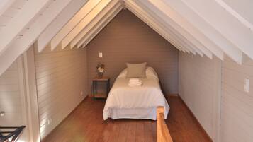 Un dormitorio con techo inclinado, suelo de madera, una cama con ropa blanca y una mesita de noche con un florero.
