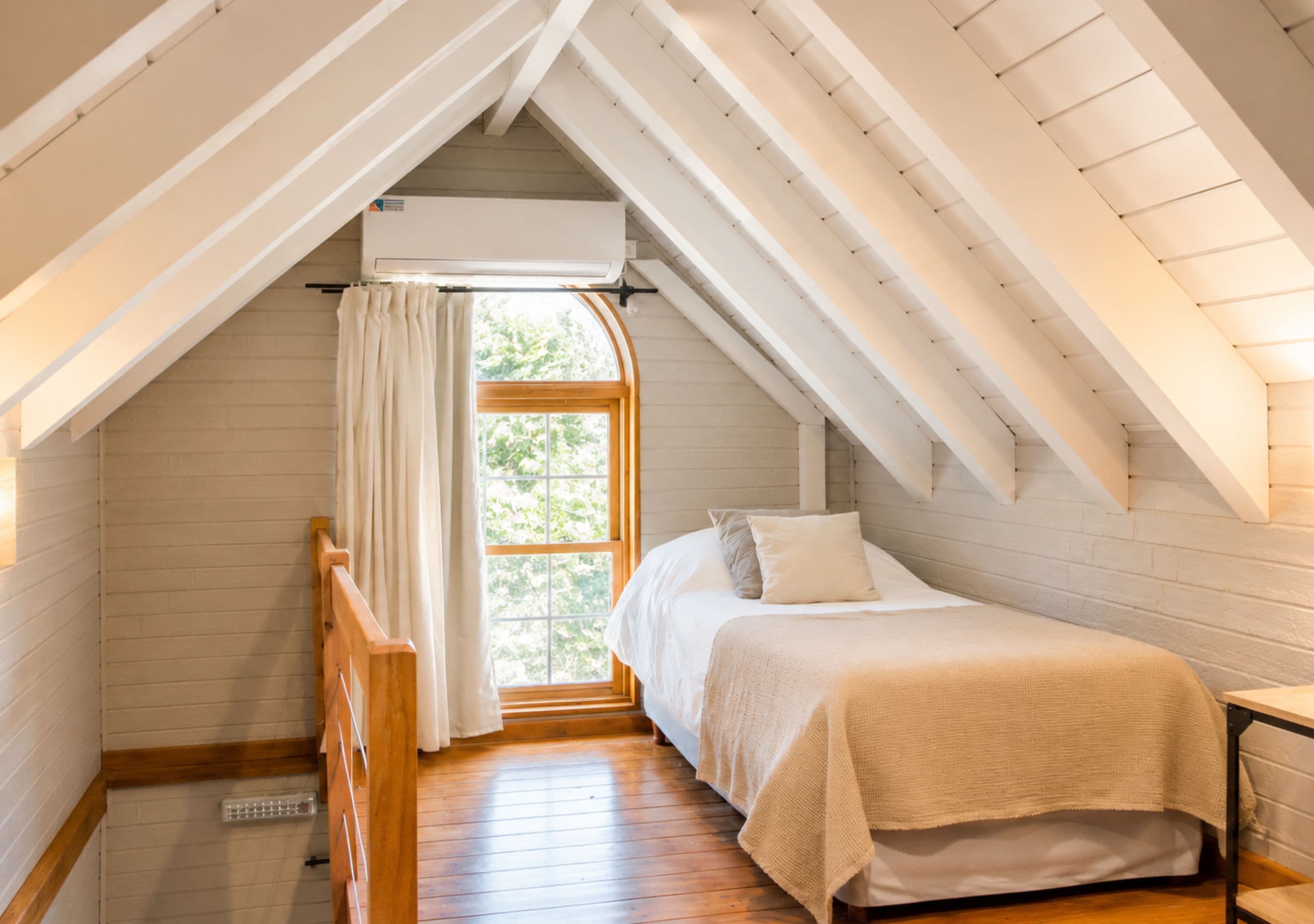 Une chambre avec un plafond en pente, une fenêtre avec des rideaux, un lit avec des oreillers et un plancher en bois.