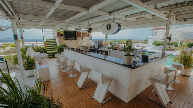 Zona de bar con vista a la playa, taburetes blancos y plantas en macetas.