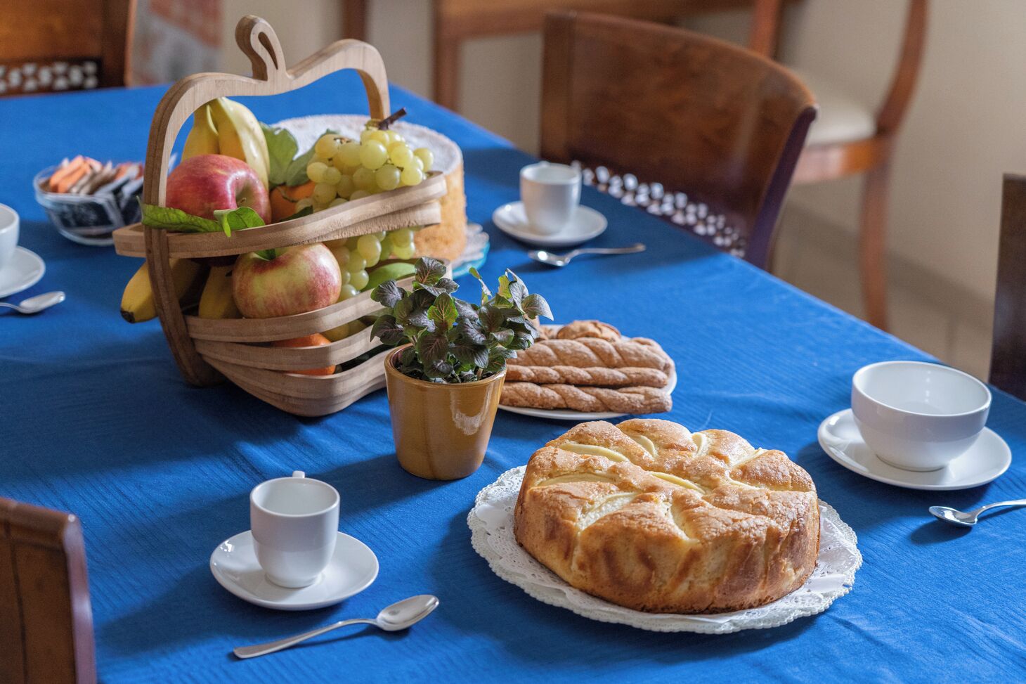 Une table dressée avec une nappe bleue, agrémentée d’un panier de fruits, d’une plante en pot, d’un pain tressé, d’une pâtisserie ronde, ainsi que de tasses et de soucoupes blanches.