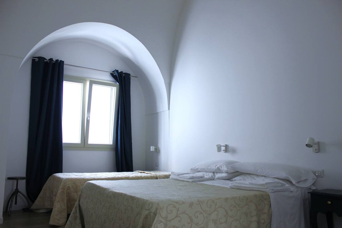A room with two beds, a window with curtains, a small table, and a wall-mounted light fixture.